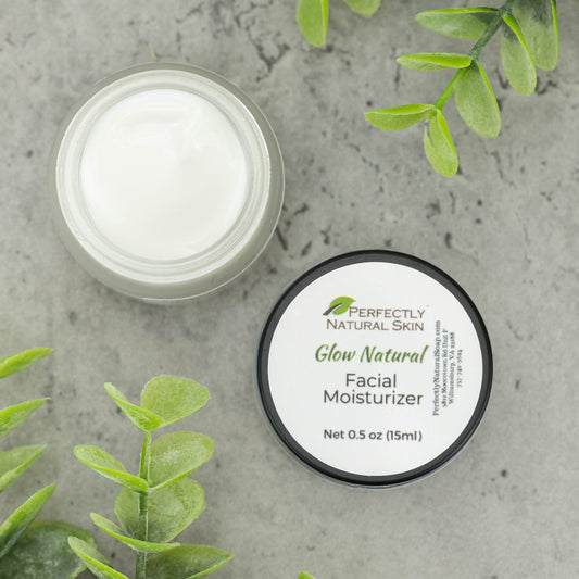 A small round jar of Perfectly Natural Skin Glow Natural Facial Moisturizer with a white label and black lid, placed on a gray stone-like surface with green leaves in the background.