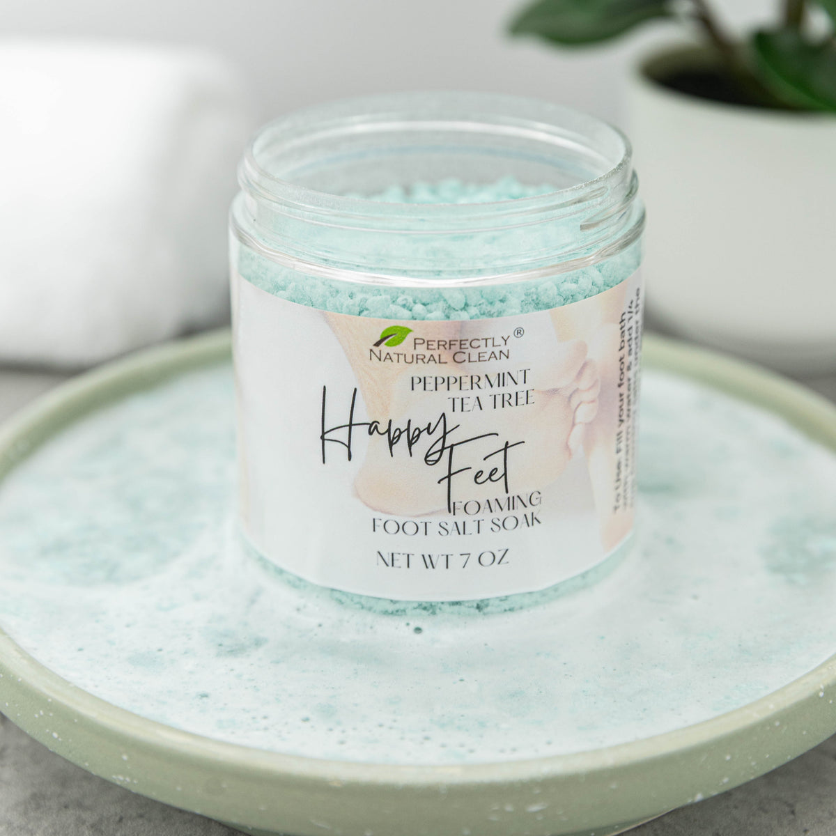 A jar of Perfectly Natural Clean Happy Feet Foaming Foot Salt Soak in Peppermint Tea Tree scent, filled with light blue salt crystals and resting on a matching light green ceramic dish.