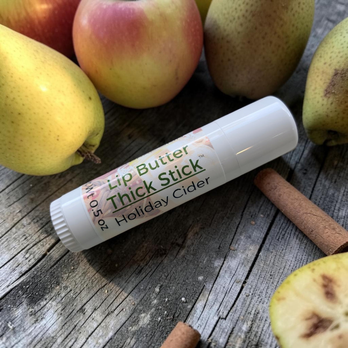 A white lip butter thick stick labeled 'Holiday Cider' lies on a rustic wooden surface surrounded by apples and cinnamon sticks.