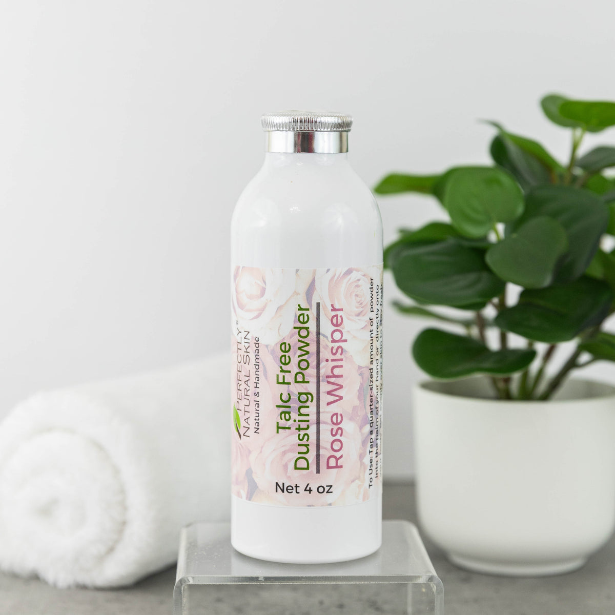 A white bottle of Perfectly Natural Skin's Talc Free Dusting Powder in Rose Whisper scent, displayed on a clear acrylic stand. The background features a soft-focus white towel on the left and a green potted plant on the right, set against a clean white backdrop.