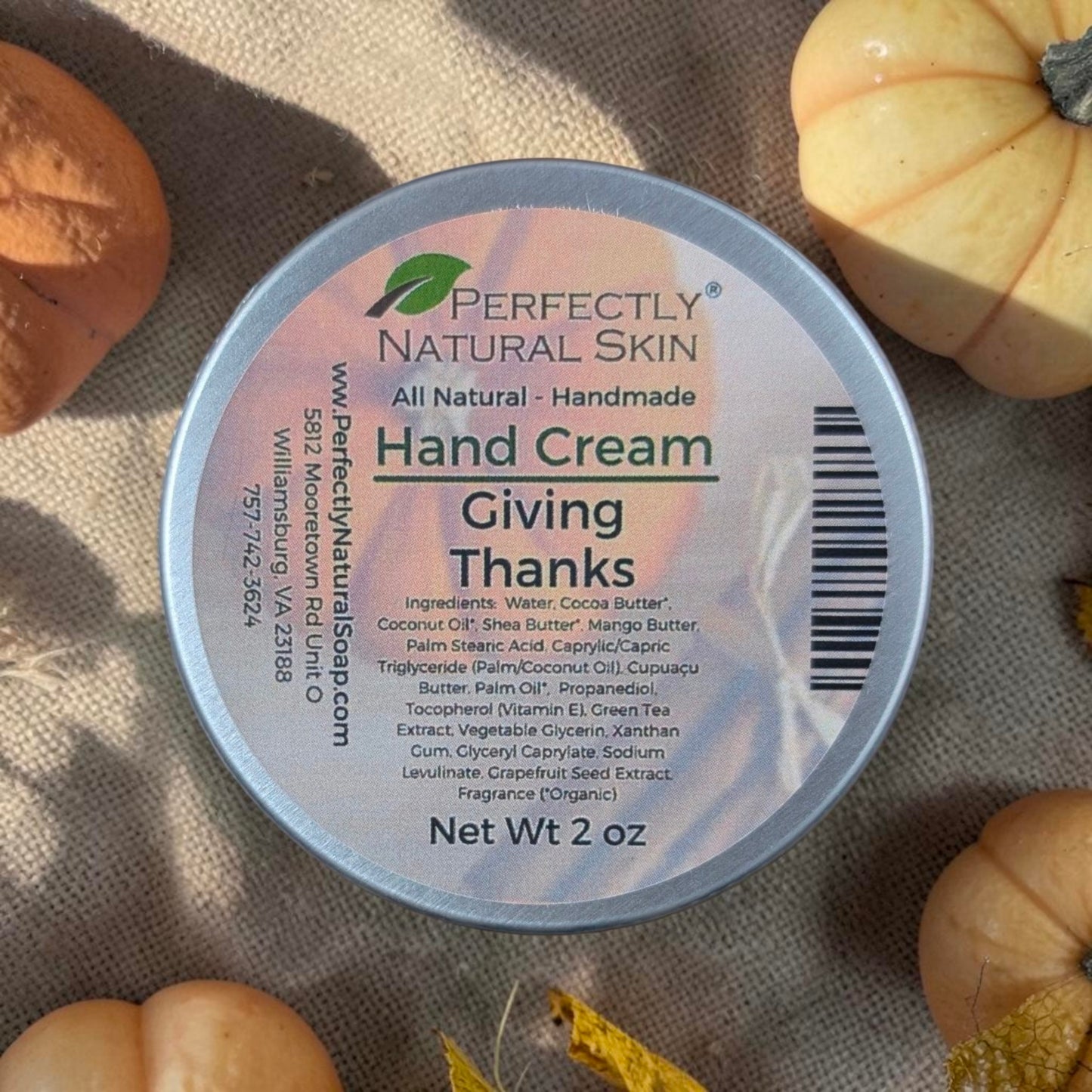 A round tin of Perfectly Natural Skin Hand Cream labeled 'Giving Thanks' sits on a textured beige fabric surrounded by small pumpkins and autumn leaves.
