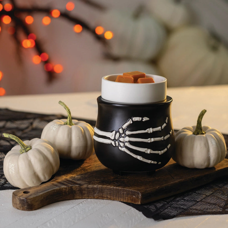 A Halloween-themed ceramic wax warmer with a skeleton hand design, holding a white cup filled with orange wax melts, styled on a wooden board with small pumpkins and black spiderweb fabric.