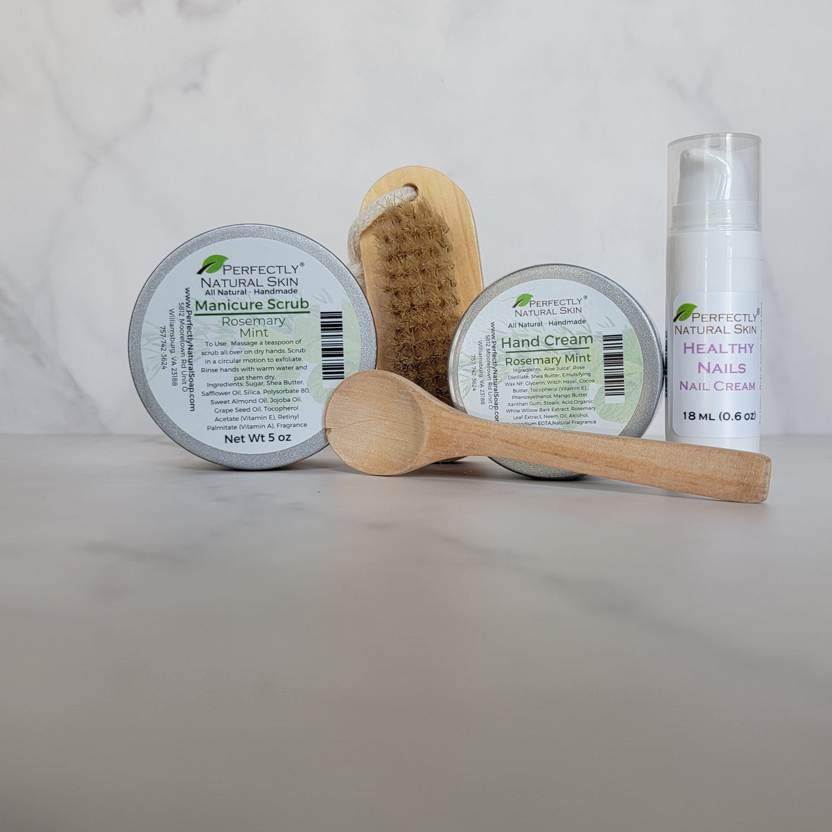 A manicure and nail care set from Perfectly Natural Skin, including a mint scrub tin, a hand cream tin, a wooden brush, a wooden spoon, and a white nail cream bottle, arranged on a light marble surface.
