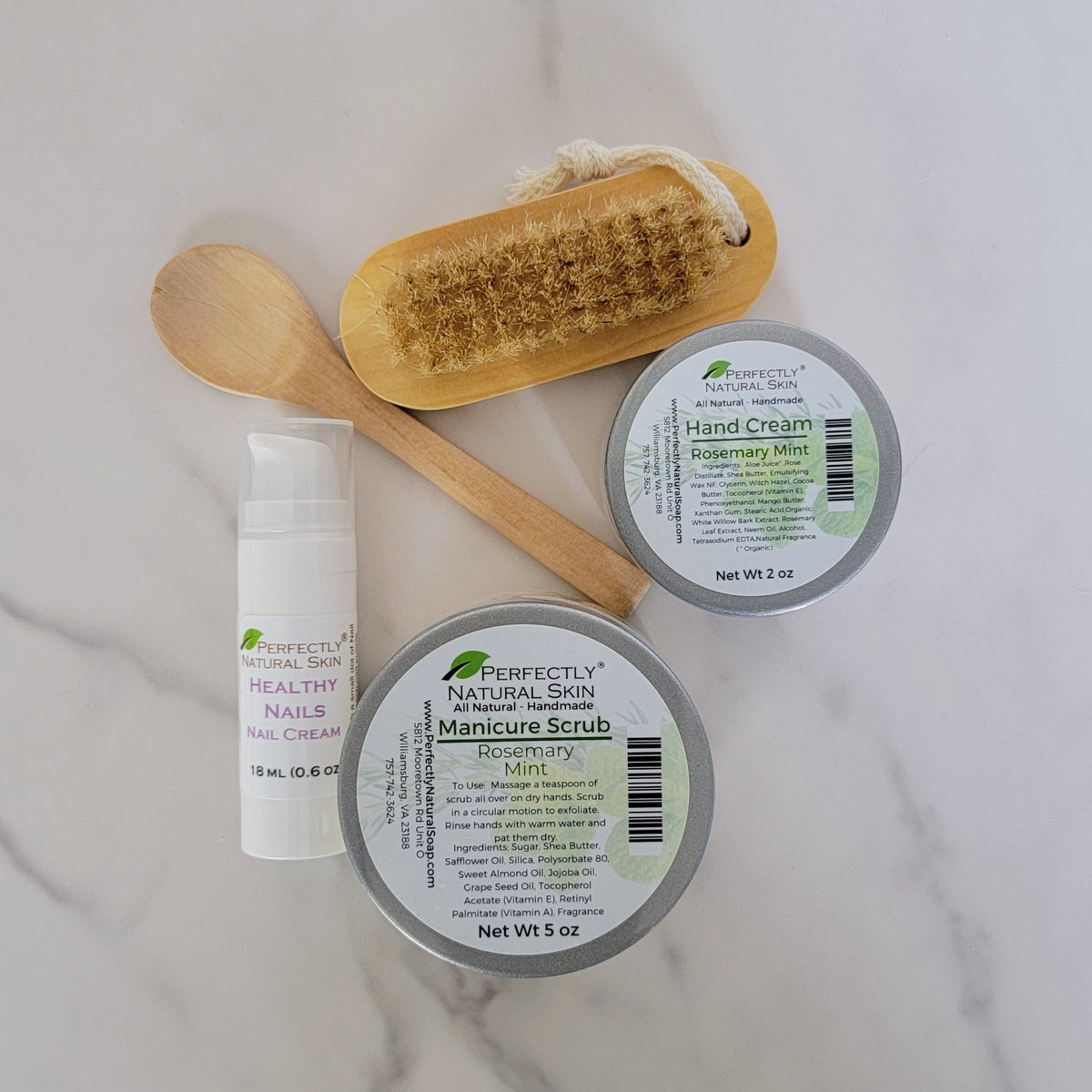A flat-lay of a manicure and nail care set on a white marble background, including a wooden scrubber, wooden spoon, tin of rosemary mint manicure scrub, hand cream, and a tube of nail cream by Perfectly Natural Skin.