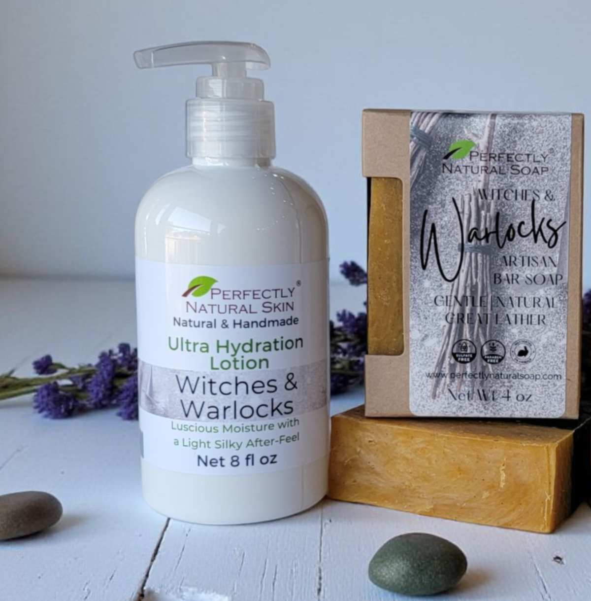 A bottle of Perfectly Natural Skin Ultra Hydration Lotion stands next to a boxed bar of Witches & Warlocks artisan bar soap on a wooden block, styled with purple flowers and smooth stones on a white wooden surface.