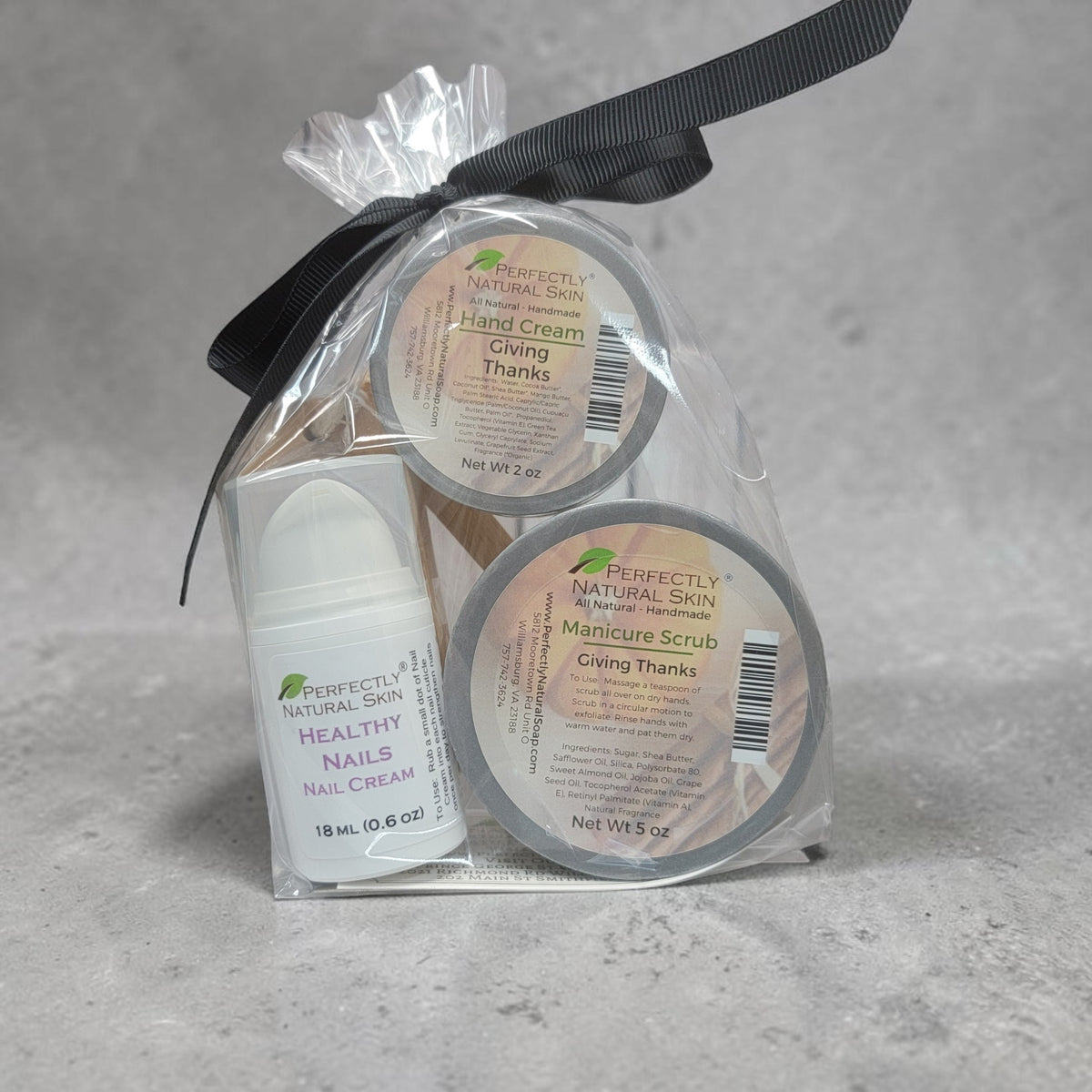 A gift set of three nail care products from Perfectly Natural Skin, including Hand Cream, Manicure Scrub, and Healthy Nails Nail Cream, packaged in a clear plastic bag tied with a black ribbon, set against a textured gray background.