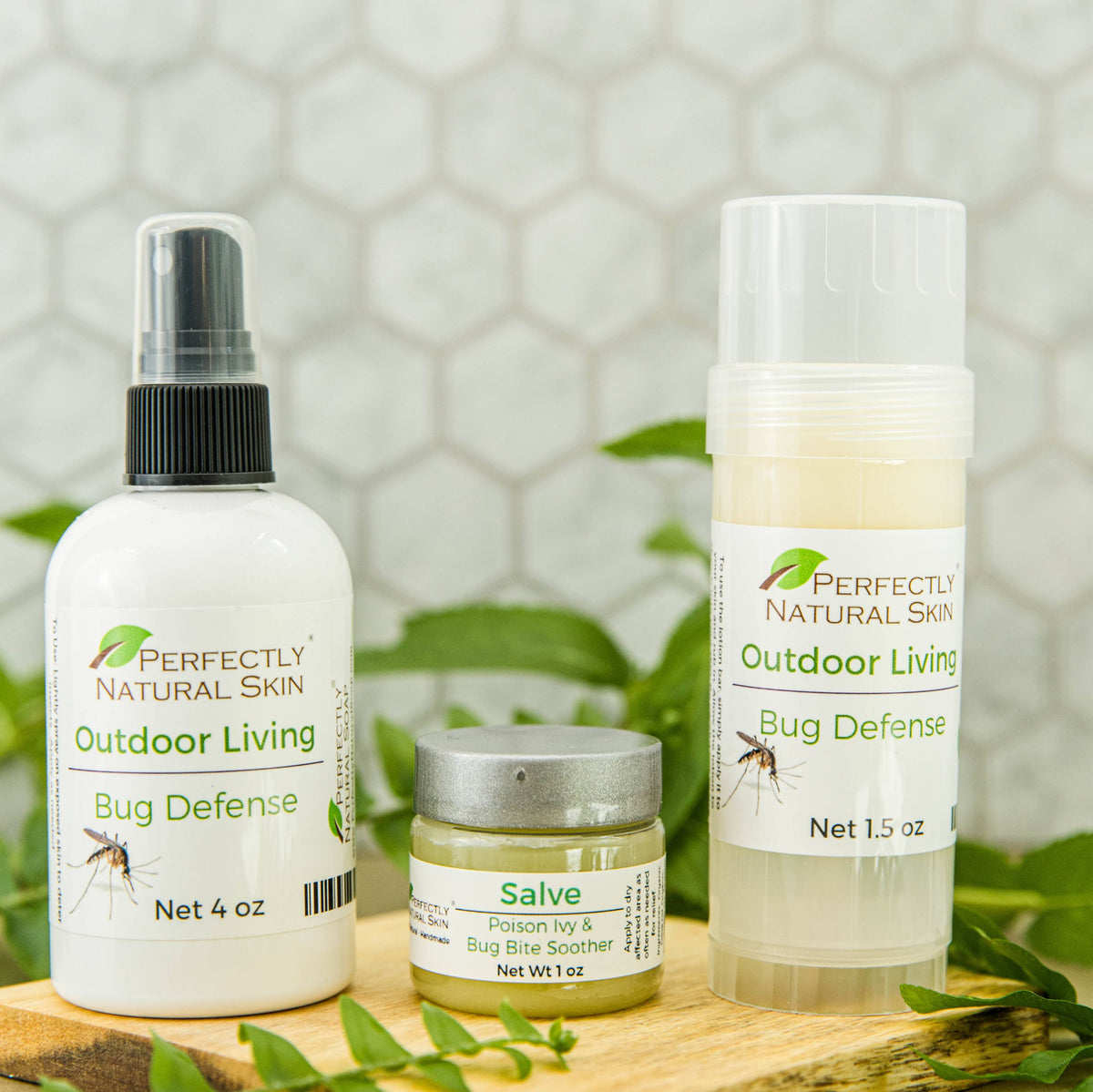 Three outdoor skin care products from Perfectly Natural Skin are displayed on a wooden surface with green leaves and a white hexagonal tile background. 