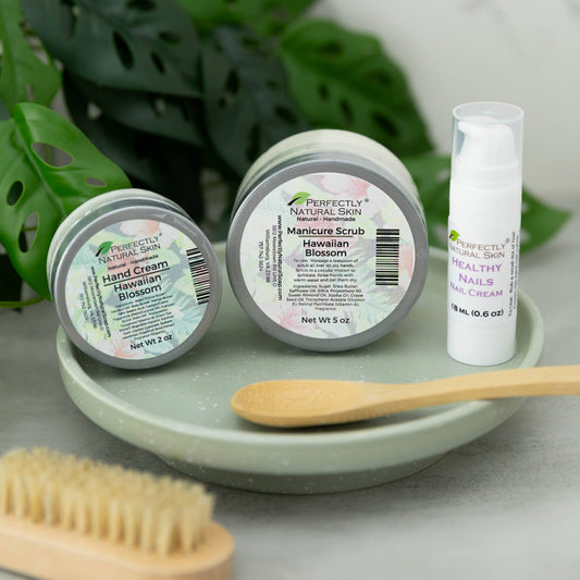 Three skincare products from Perfectly Natural Skin are arranged on a light green ceramic tray: a hand cream jar, a manicure scrub jar, and a healthy nails cream tube. A wooden spoon and brush rest in front, with large green leaves in the background.