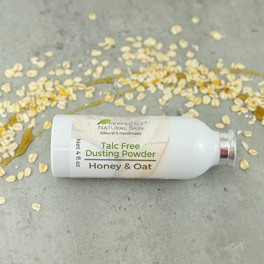 A white bottle of Perfectly Natural Skin Honey & Oat Talc Free Dusting Powder lies on its side on a gray concrete surface, surrounded by scattered oats and a drizzle of honey.