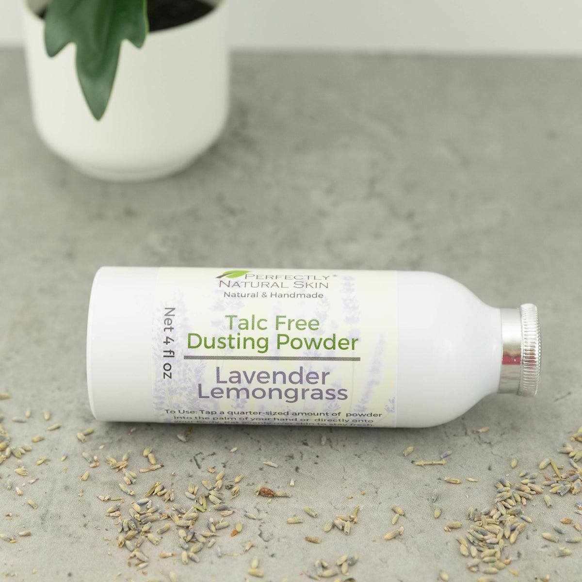 A white bottle of Perfectly Natural Skin Lavender Lemongrass Talc Free Dusting Powder lies on a gray stone surface with scattered dried lavender buds around it and a blurred potted plant in the background.