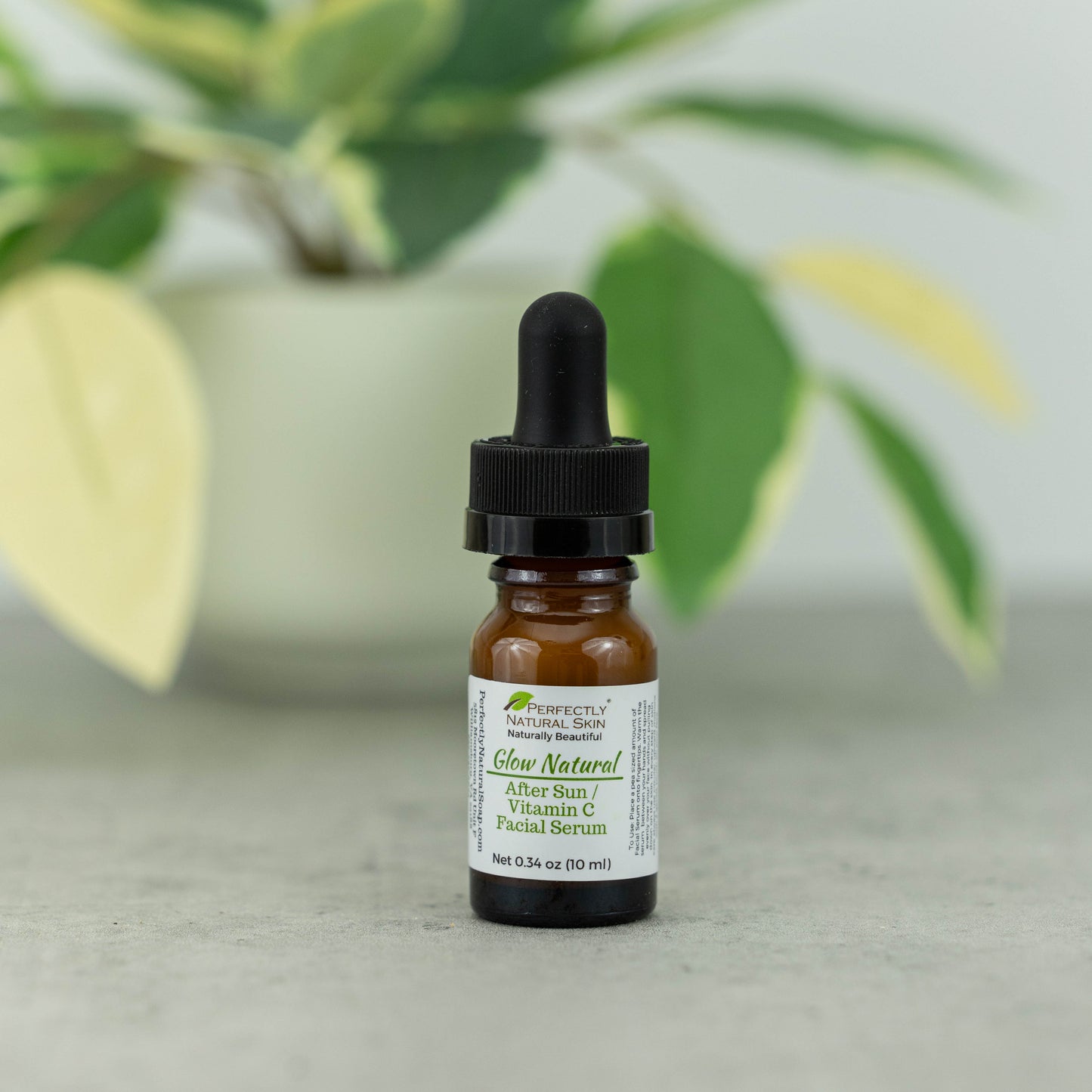 A small amber glass dropper bottle of Perfect's Natural Skin Glow Natural Algae Sun / Vitamin C Facial Serum, standing on a light gray surface with a blurred green plant in the background.