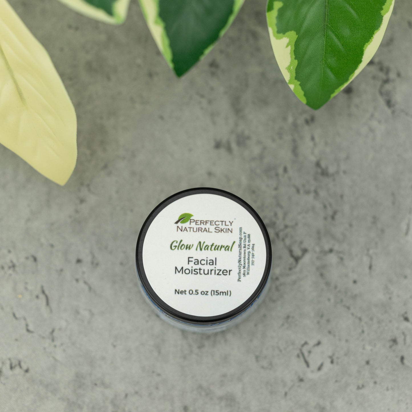 A top-down view of a round black jar of Perfectly Natural Skin Glow Natural Facial Moisturizer, placed on a gray stone surface with green and yellow variegated leaves in the background.