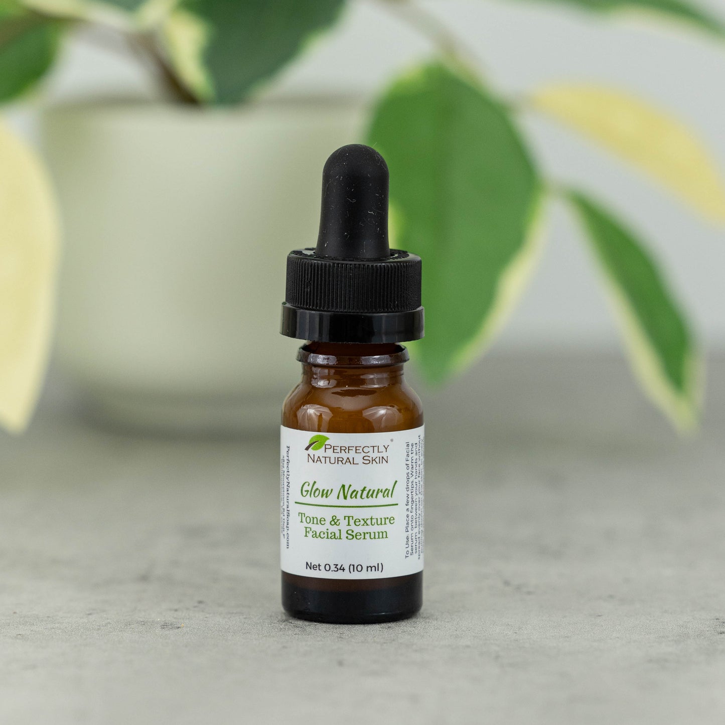 A small amber glass dropper bottle of Perfectly Natural Skin Glow Natural Tone & Texture Facial Serum, standing on a gray surface with a blurred green plant in the background.