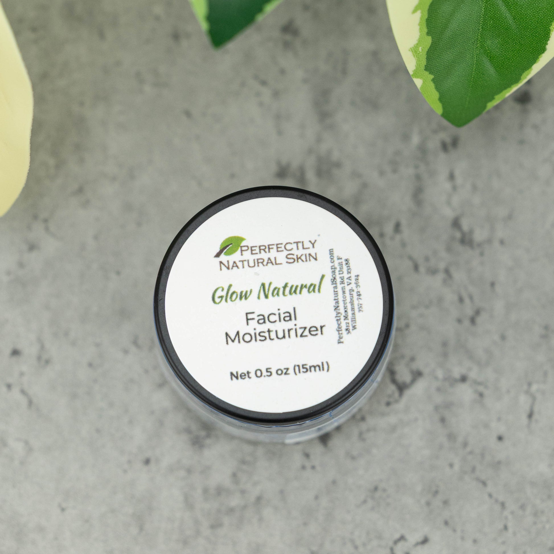 A small round jar of Perfectly Natural Skin Glow Natural Facial Moisturizer with a white label and black lid, placed on a gray stone-like surface with green leaves in the background.