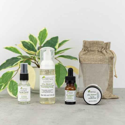 A skincare gift set from Perfectly Natural Skin arranged on a gray surface with a potted plant in the background. The set includes a facial toner, a foaming soda spray, an oil bottle with a dropper, a jar of facial moisturizer, and a burlap drawstring pouch.