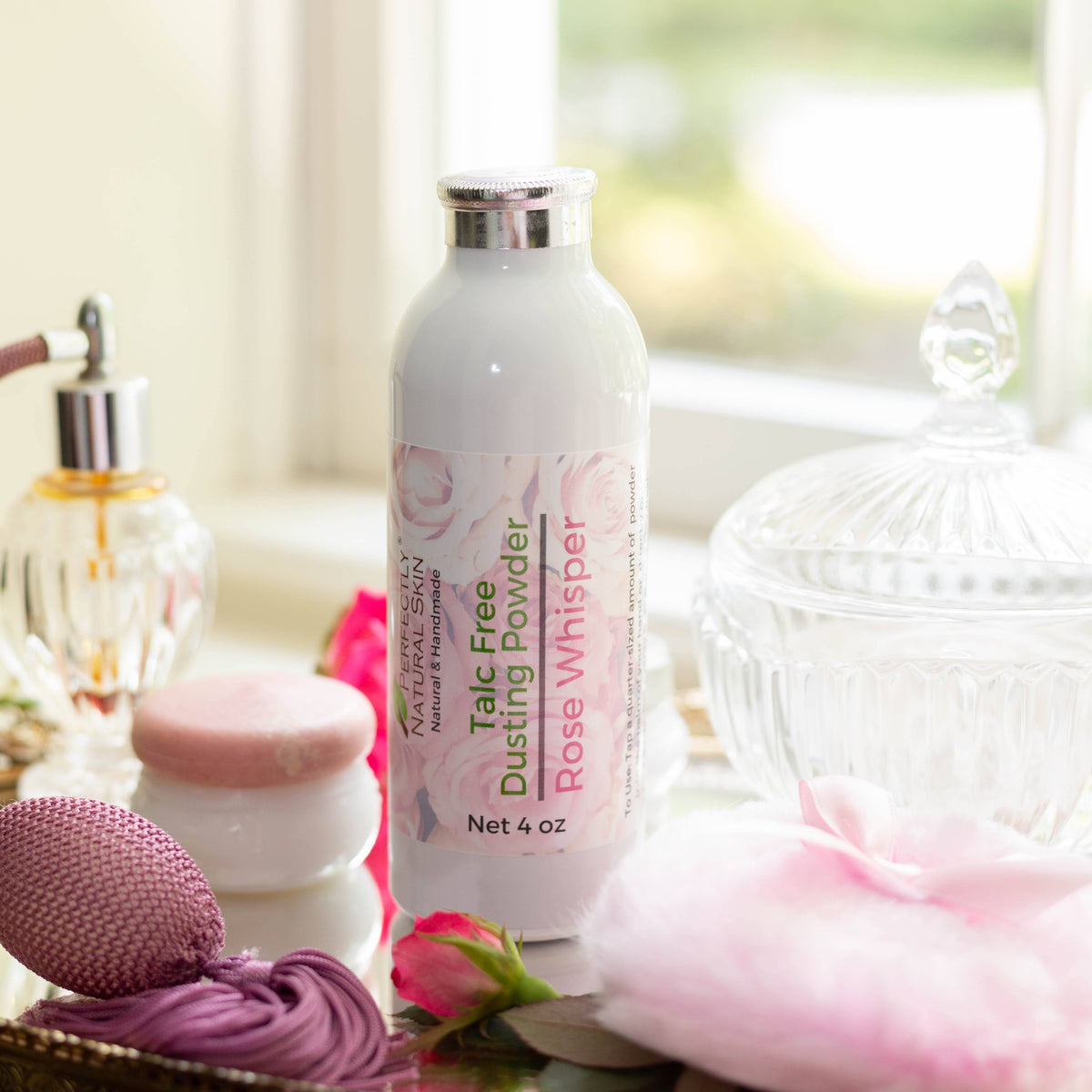 Talc Free Dusting Powder in Rose Whisper scent by Perfectly Natural Soap, styled on a tabletop with pink rosebuds and a powder puff