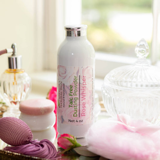 A bottle of Talc Free Dusting Powder Rose Whisper by Kari's Natural & Beautiful Skincare, styled on a tabletop with pink rosebuds, a pink sponge, and a glass jar in a bright, soft-focus setting.