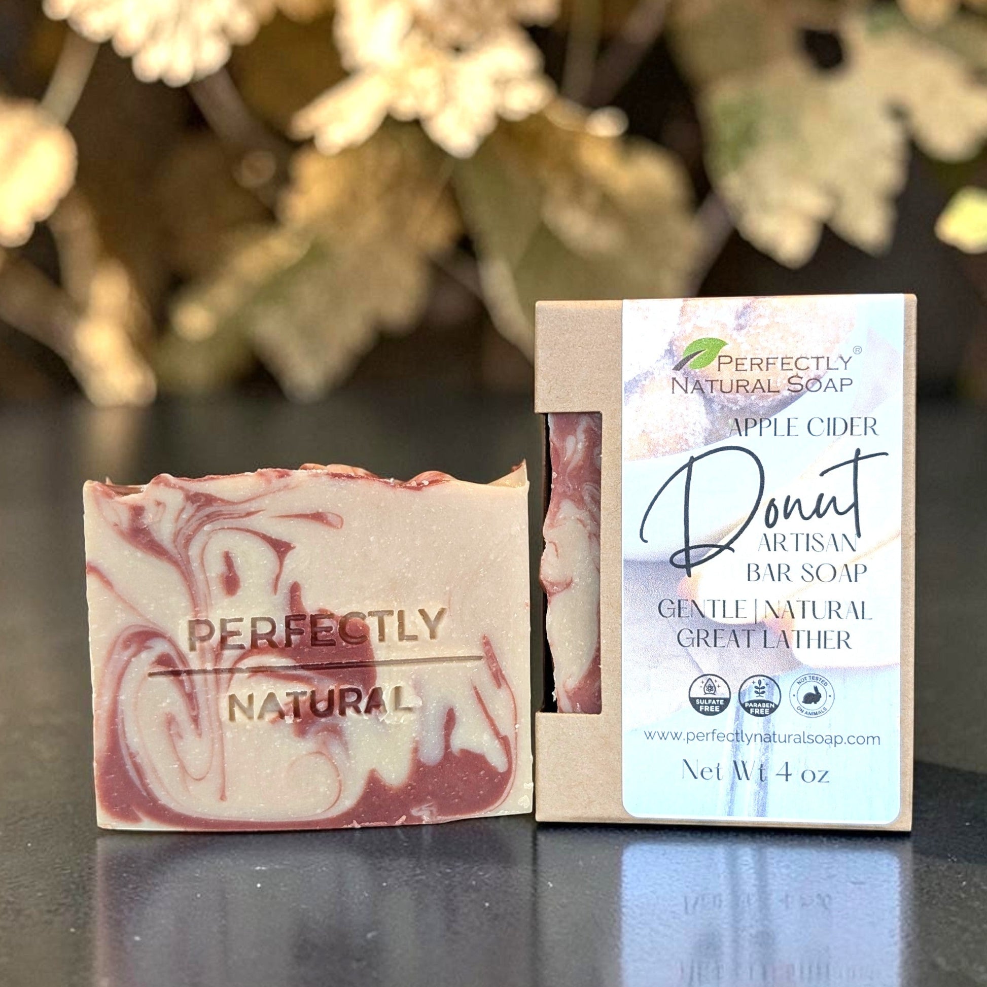 A bar of marbled apple cider donut natural bar soap is shown next to its paper packaging box on a dark reflective surface with a blurred natural background.