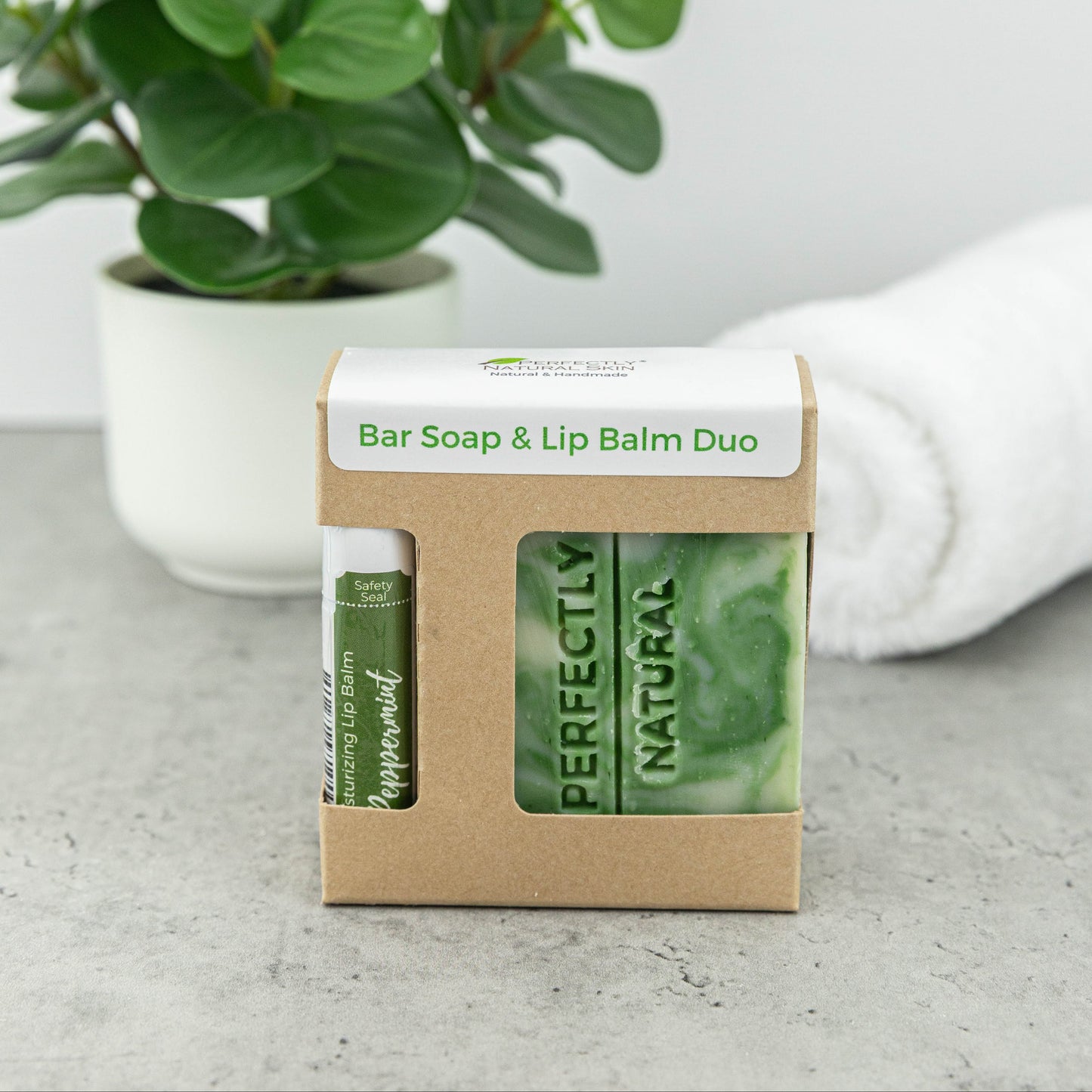 A bar soap and lip balm duo in a kraft paper box, displayed on a gray surface with a potted green plant and a white towel in the background.