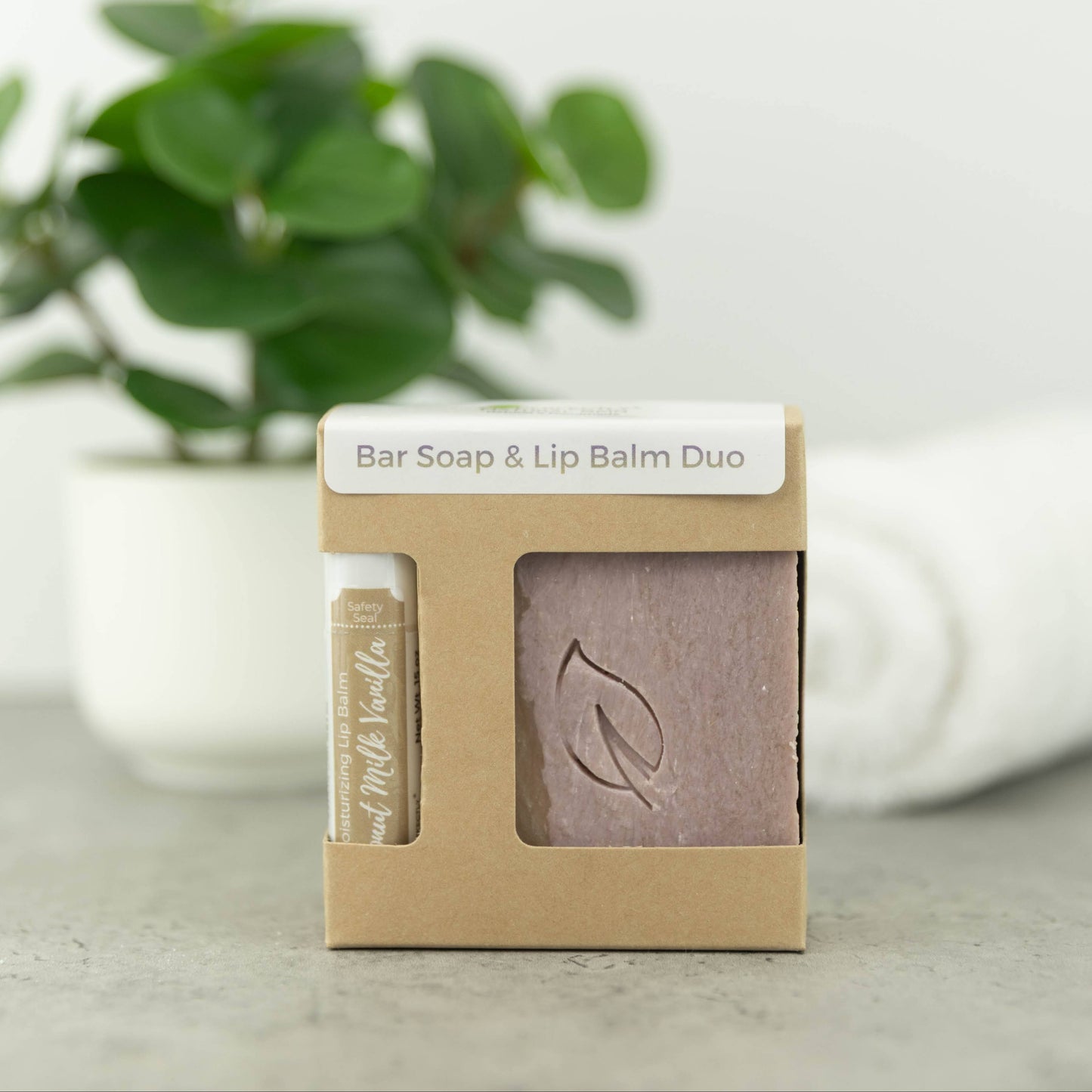 A bar soap and lip balm duo in a small kraft paper box, with a visible bar of soap and a lip balm tube inside. The box is labeled 'Bar Soap & Lip Balm Duo' and sits on a light surface with a blurred green plant and white towel in the background.