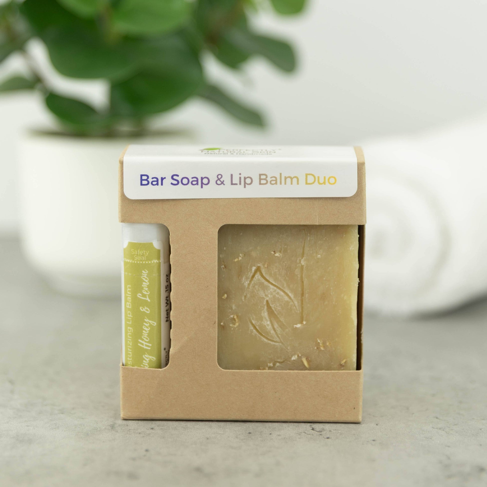 A bar soap and lip balm duo in a small kraft paper box with a clear window, shown on a light surface with a blurred green plant in the background.