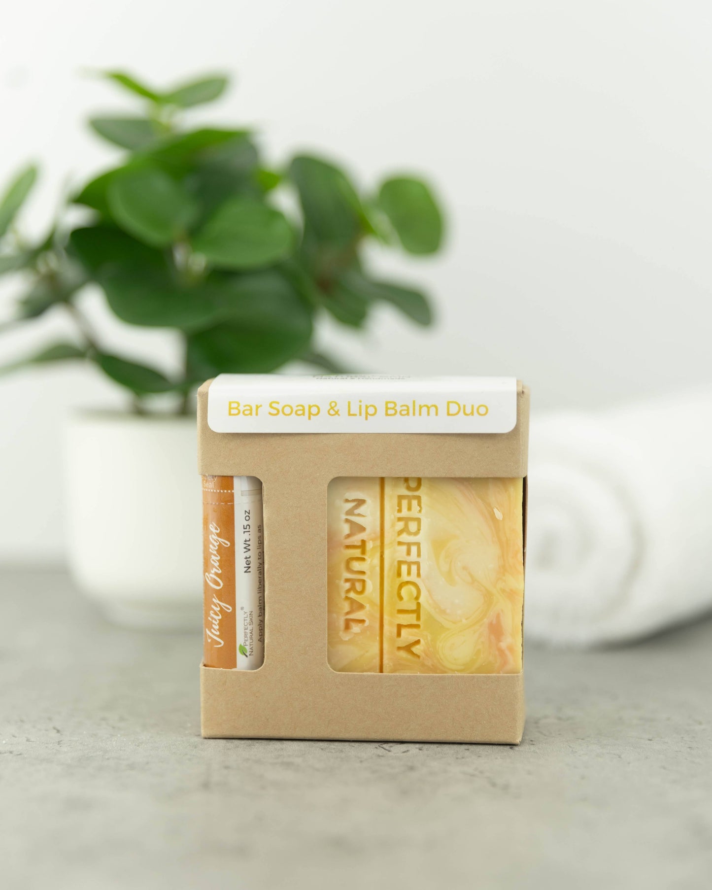 A bar soap and lip balm duo in a small kraft paper box with a white label reading 'Bar Soap & Lip Balm Duo.' The box contains a round balm tube and a rectangular bar of soap with yellow-orange marbling.