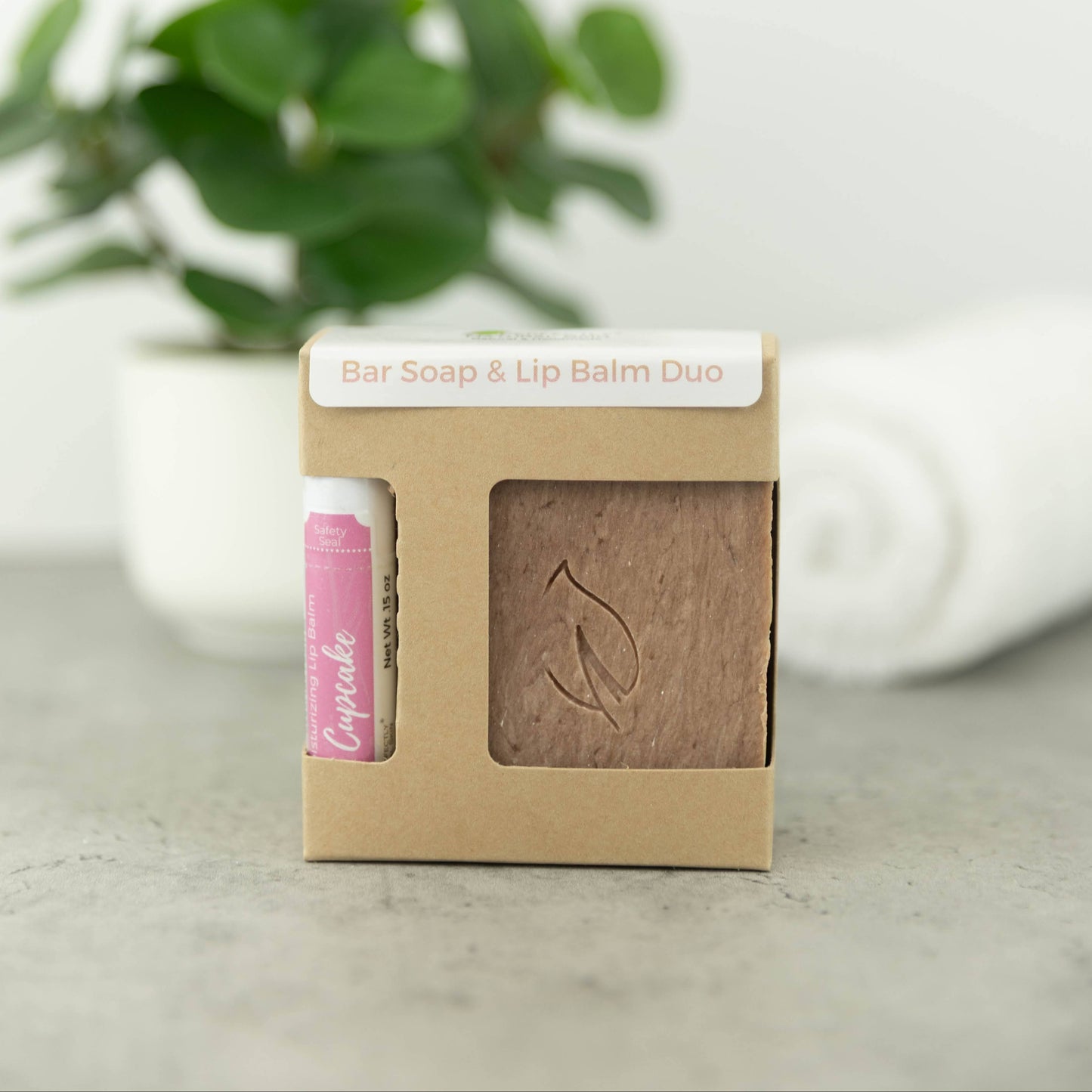 A bar soap and lip balm gift set in a kraft paper box, with a pink lip balm tube visible on the left and a brown bar of soap on the right. The box is labeled 'Bar Soap & Lip Balm Duo' and is set against a soft-focus background with a potted green plant and a white towel.