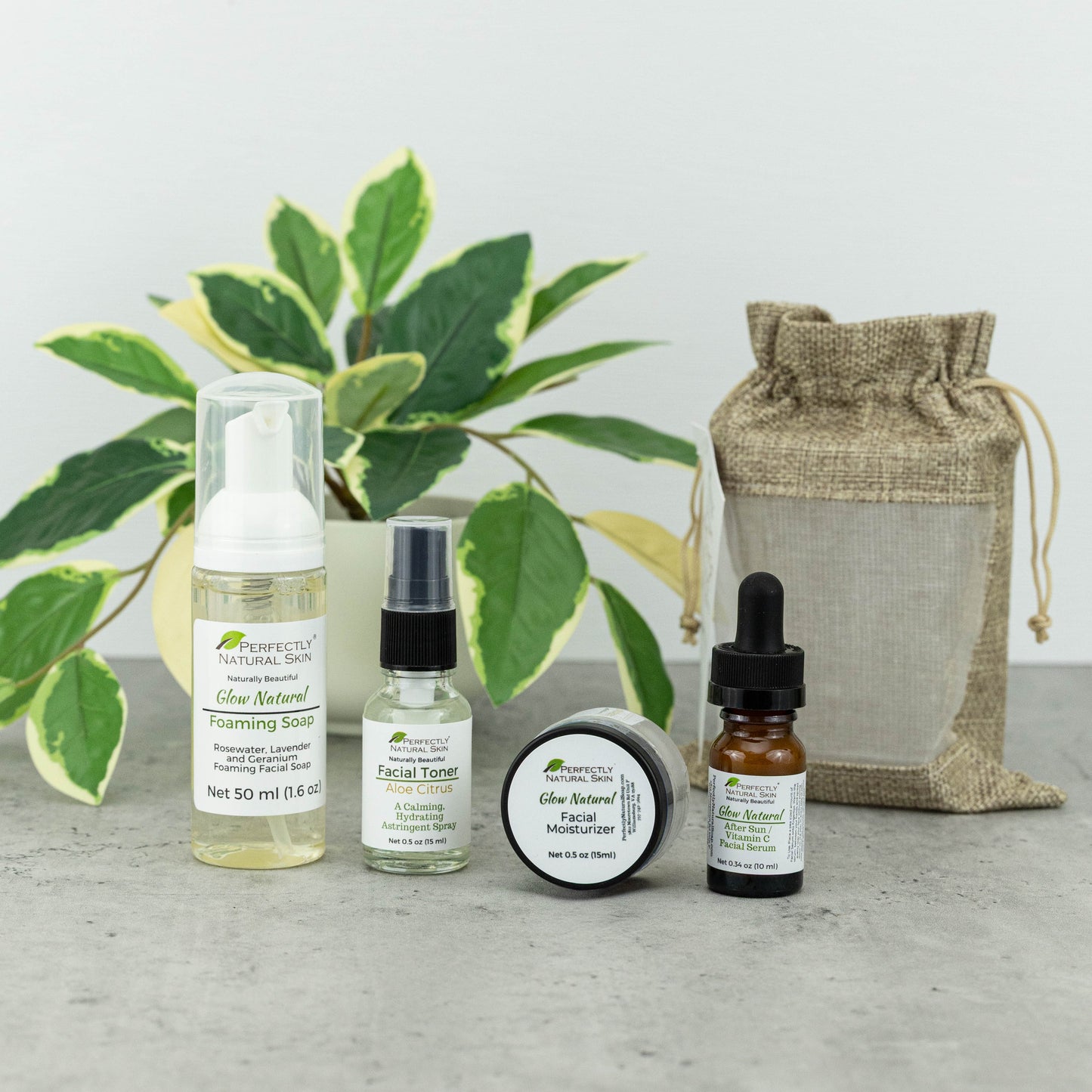 A skincare gift set from Perfectly Natural Skin arranged on a gray surface in front of a potted variegated plant. The set includes Foaming Soap, Facial Toner, a round jar of Glow Natural Facial Moisturizer, and an amber dropper bottle, alongside a burlap drawstring pouch.