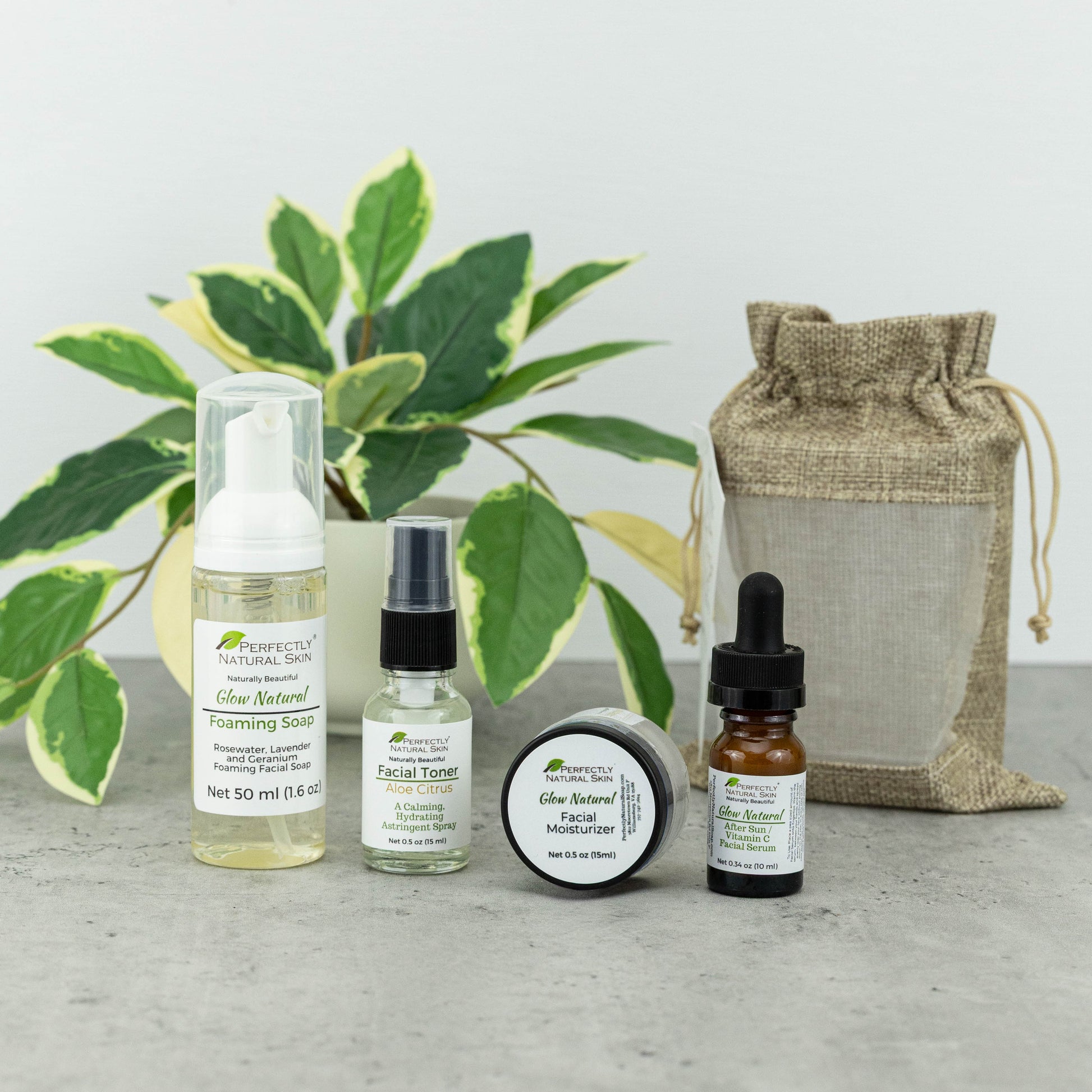 A skincare gift set from Perfectly Natural Skin arranged on a gray surface in front of a potted variegated plant. The set includes Foaming Soap, Facial Toner, a round jar of Glow Natural Facial Moisturizer, and an amber dropper bottle, alongside a burlap drawstring pouch.