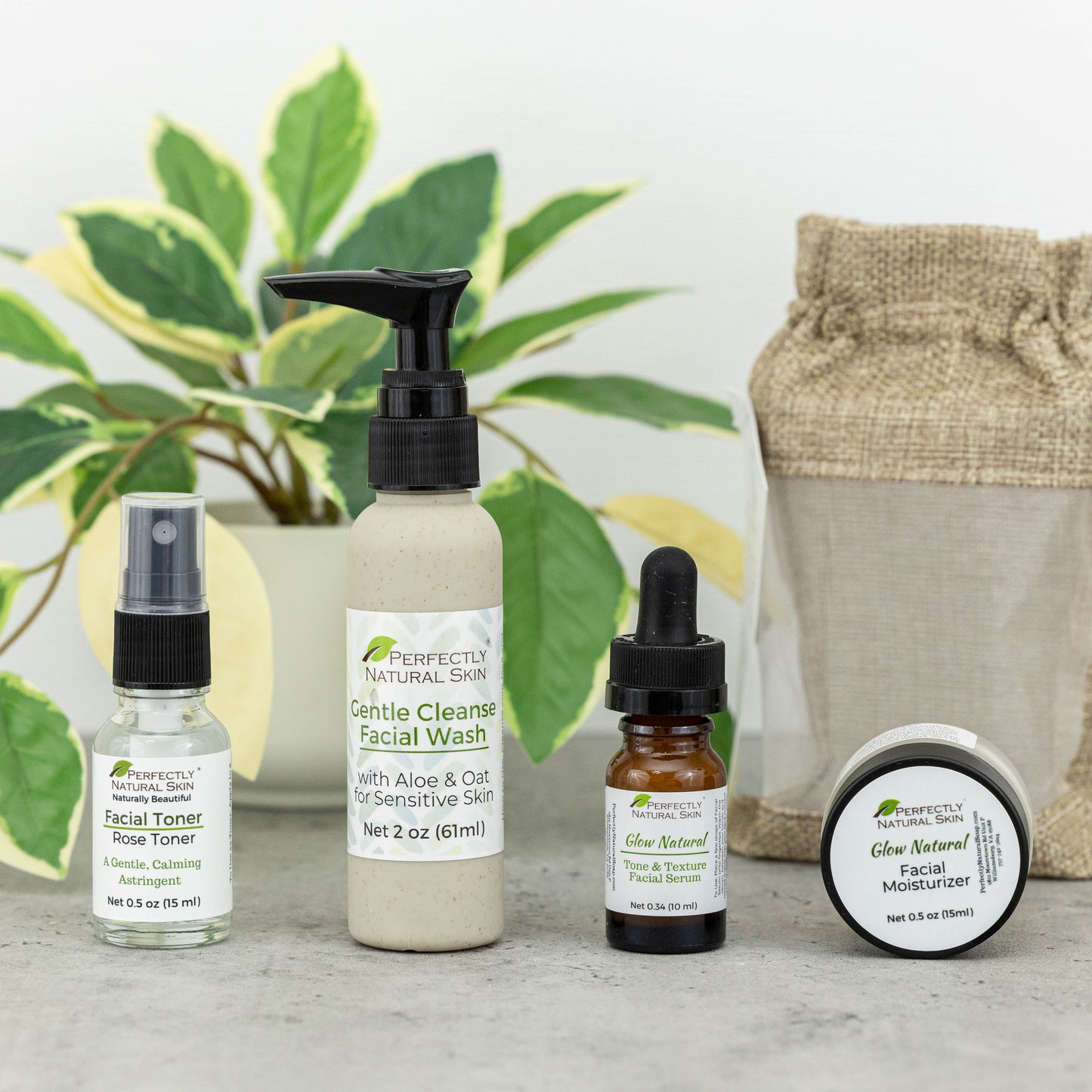 A skincare gift set from Perfectly Natural Skin arranged on a light gray surface with a potted green plant and a burlap drawstring bag in the background. The set includes a facial toner spray, a large facial cleanser pump bottle, a small dropper bottle, and a round facial moisturizer jar.