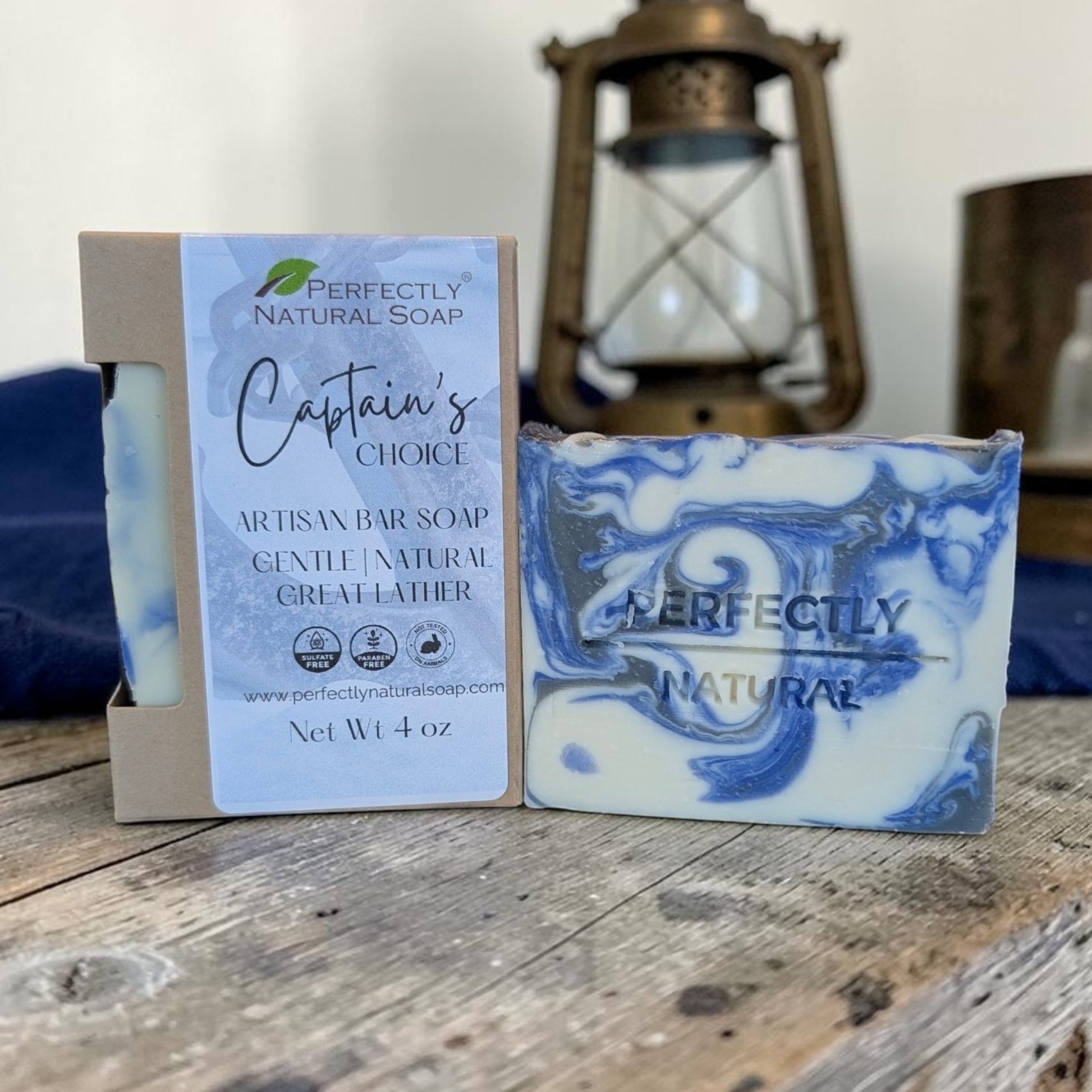 A bar of soap and its packaging box displayed on a rustic wooden table, with a blurred lantern in the background. The soap is blue and white marbled, and the box has a light blue label reading 'Perfectly Natural Soap Captain's Choice'.