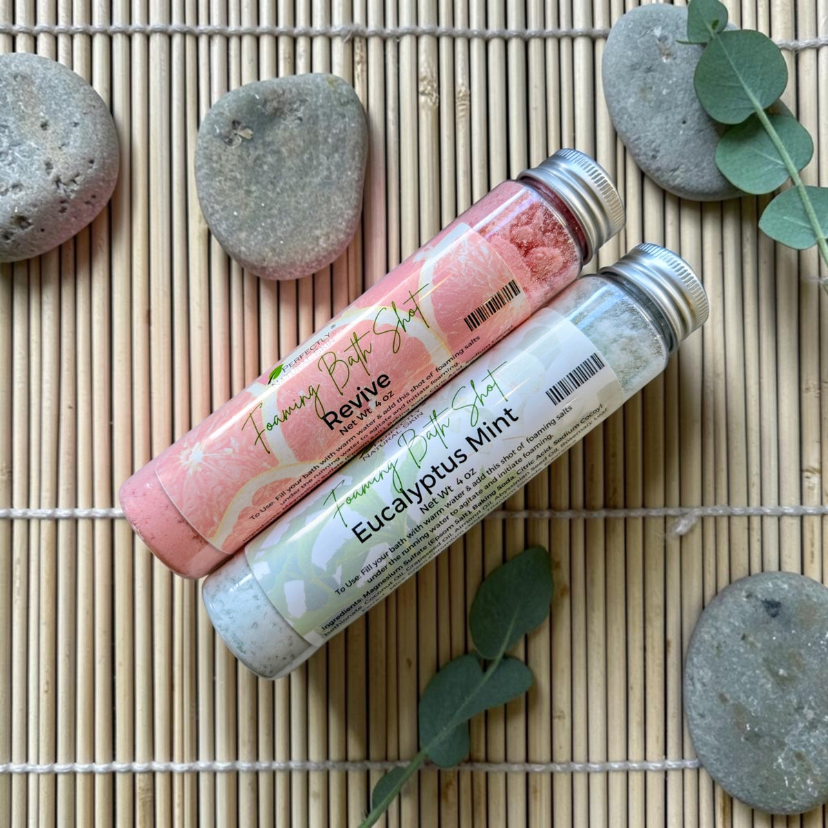 Two tubes of foaming bath shot powder, one pink labeled 'Revive' and one white labeled 'Eucalyptus Mint', arranged on a bamboo mat with decorative stones and green leaves.