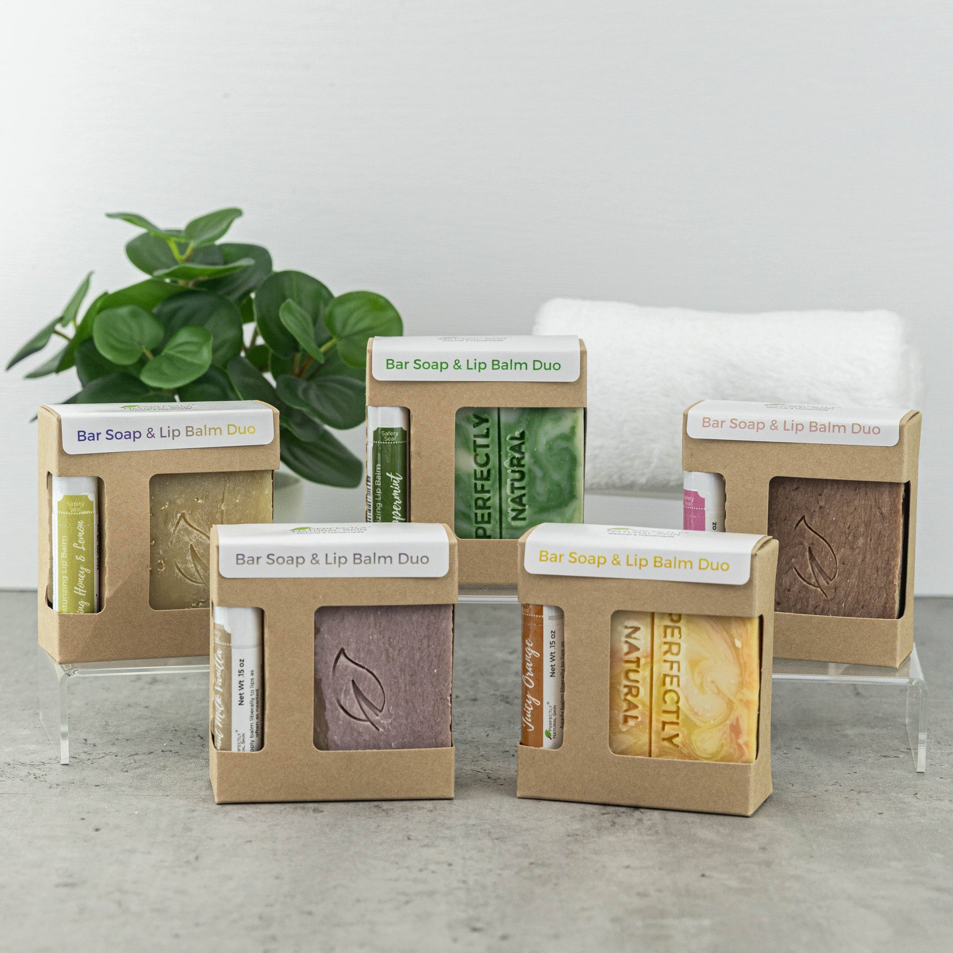 Four bar soap and lip balm duos in clear acrylic display stands, each in its own kraft paper window box. A green potted plant and a white towel are in the background against a light gray wall.