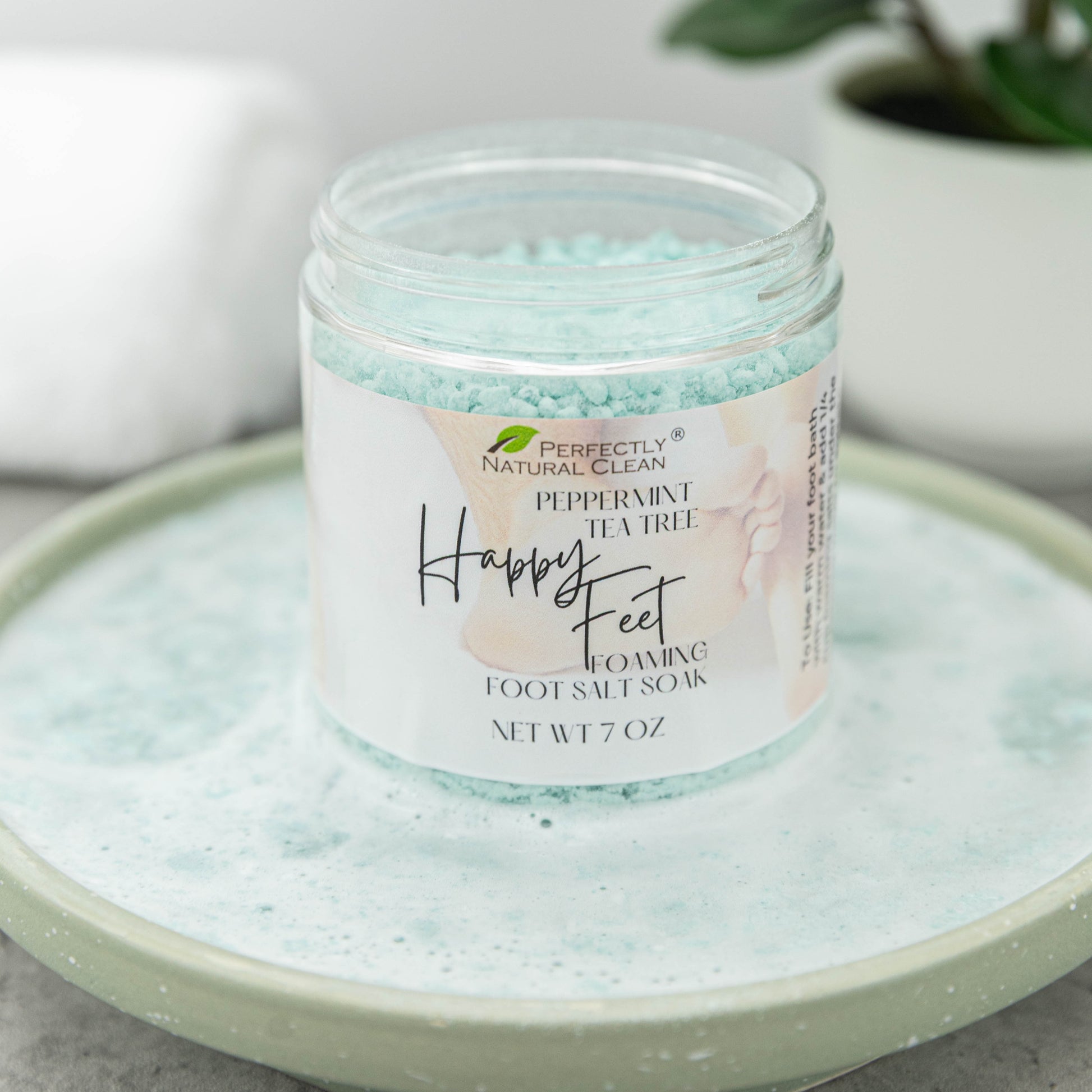 A jar of Perfectly Natural Clean Happy Feet Foaming Foot Salt Soak in Peppermint Tea Tree scent, filled with light blue salt crystals and resting on a matching light green ceramic dish.