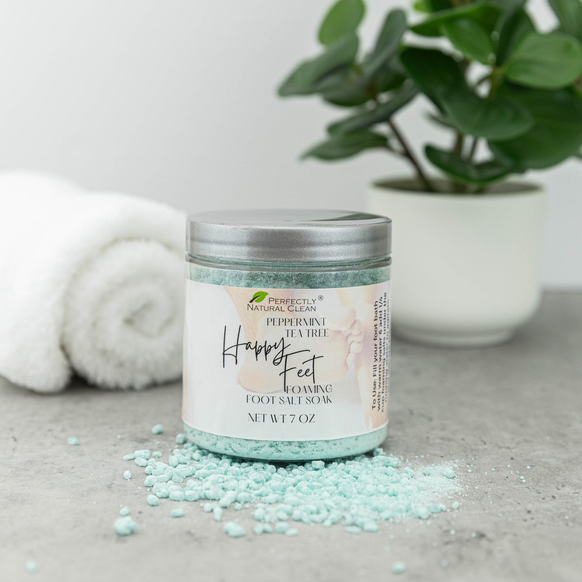 A jar of Perfectly Natural Clean Happy Feet Foaming Foot Salt Soak in Peppermint Tea Tree scent, filled with light blue-green salt crystals and topped with a silver lid, set on a gray surface with scattered crystals, a white towel, and a green plant in the background.
