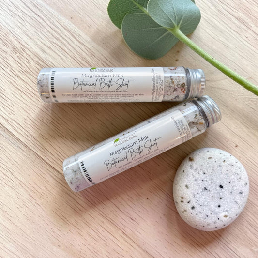 Two tubes of Perfectly Natural Soap's Botanical Bath Shot labeled Magnesium Milk Botanical Bath Shot, arranged on a light wood surface with a green leaf and a speckled stone nearby.