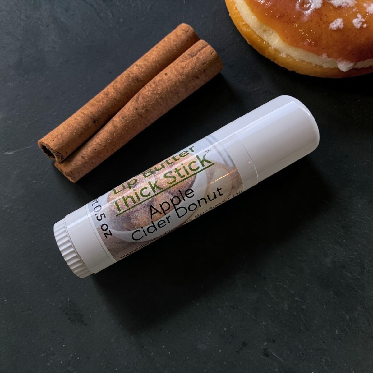 A white lip butter thick stick labeled Apple Cider Donut flavor, placed on a dark slate surface with two cinnamon sticks and part of a glazed donut visible in the background.