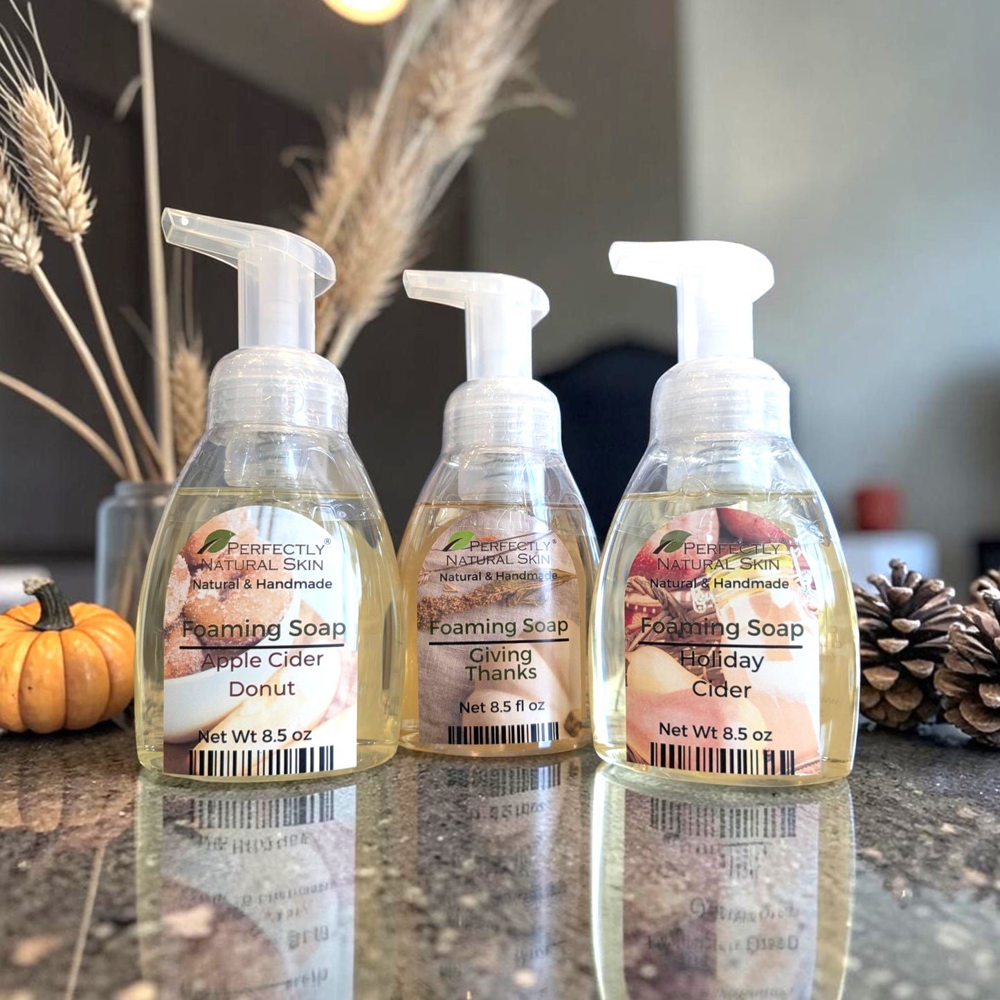 Three bottles of Perfectly Natural Skin foaming soap lined up on a reflective countertop, with dried wheat, a small pumpkin, and pinecones in the background.