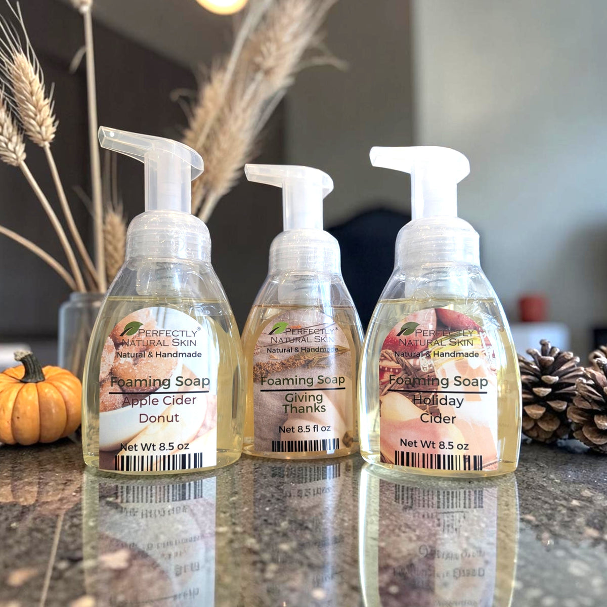 Three bottles of Perfectly Natural Skin foaming soap lined up on a reflective countertop, with dried wheat, a small pumpkin, and pinecones in the background.
