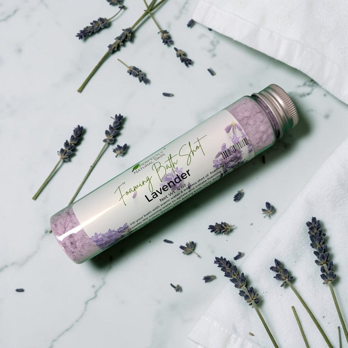 A clear cylindrical bottle of lavender foaming bath shot with a silver cap, placed diagonally on a white marble surface with scattered dried lavender sprigs and white fabric.