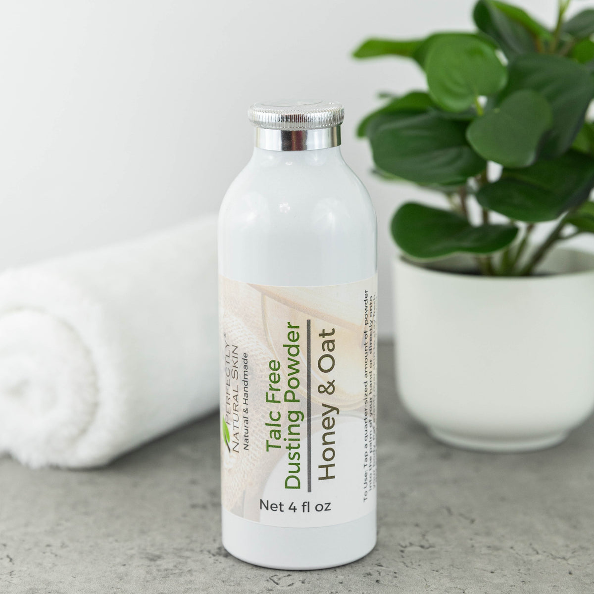 A white bottle of Perfectly Natural Skin Talc Free Dusting Powder with Honey & Oat label, set on a gray surface with a white towel and a potted green plant in the background.