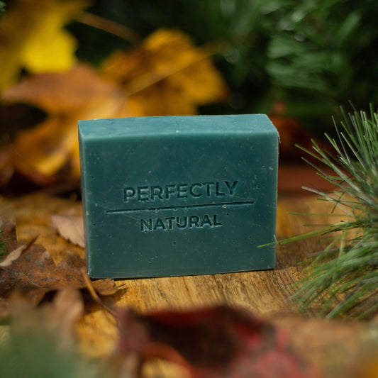 Green bar of soap labeled 'Perfectly Natural' on a wooden surface with foliage in the background
