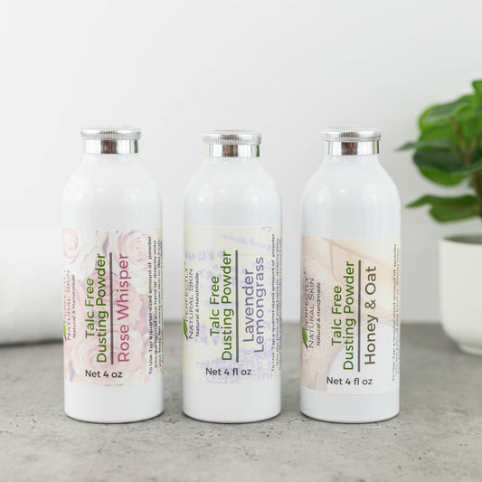 Three white bottles of Talc Free Dusting Powder lined up on a gray surface against a white background. The bottles are labeled Rose Whisper, Lemongrass, and Honey & Oat, with a small green plant visible in the background on the right.