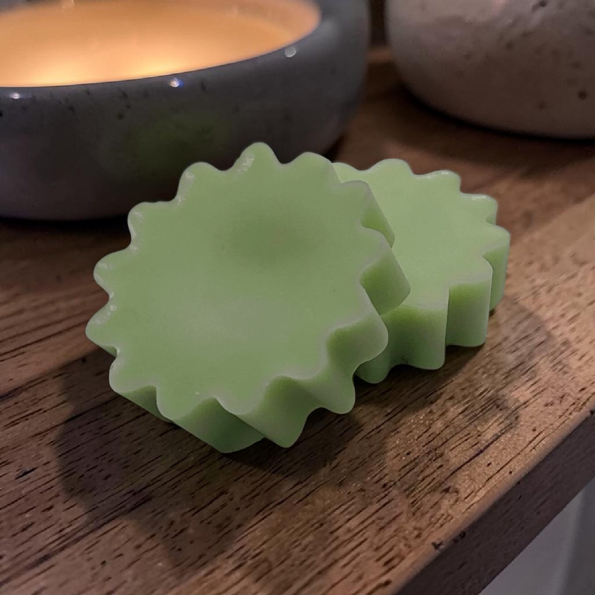 Two green wax tart molds on a wooden surface with a bowl in the background.