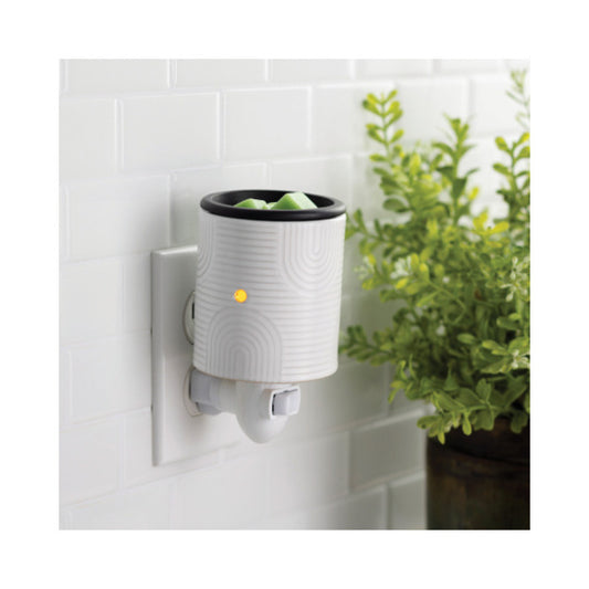 A white wax warmer with a black rim and a small yellow indicator light, mounted on a white tiled wall next to a green potted plant.