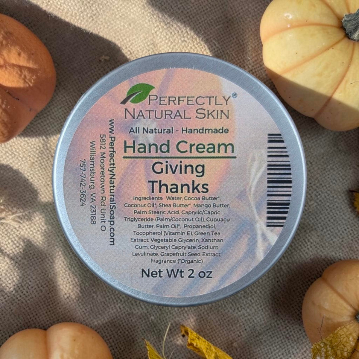 A round tin of Perfectly Natural Skin Hand Cream labeled 'Giving Thanks' sits on a textured beige fabric surrounded by small pumpkins and autumn leaves.